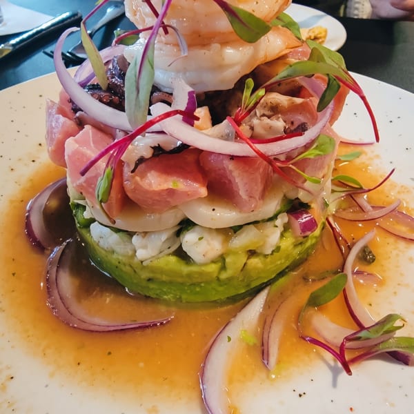 Stacked seafood ceviche with shrimp, tuna, avocado, red onion, and microgreens on a white plate.