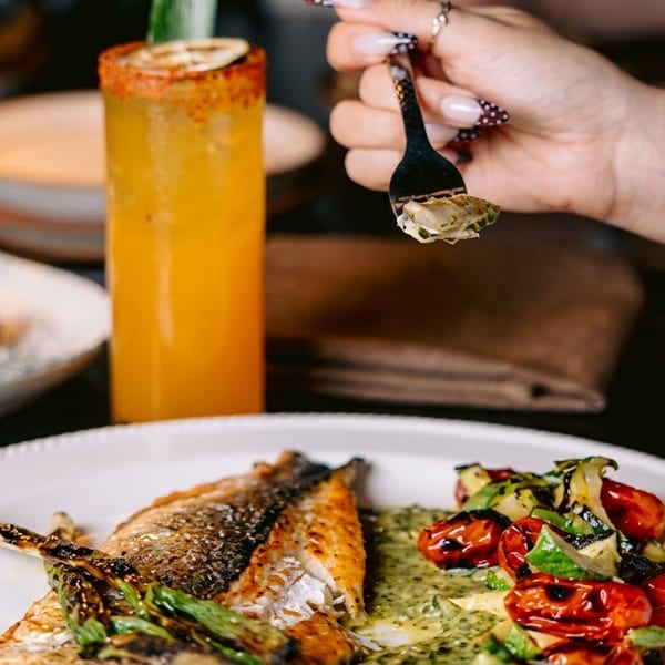 A person eating grilled fish with roasted vegetables, a cocktail, and dessert on a table.