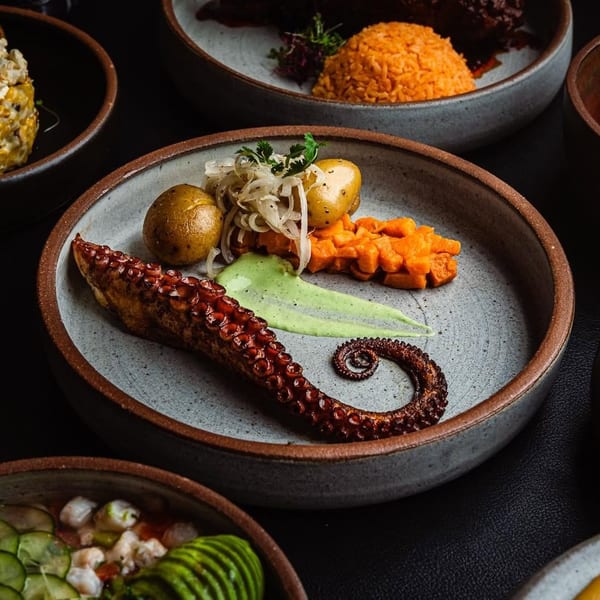 Grilled octopus with potatoes, sweet potato, and green sauce, surrounded by other dishes.
