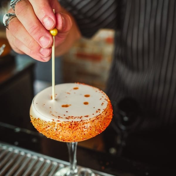 Bartender garnishing a cocktail with white foam, chili-dusted rim, and a golden bead on a pick.