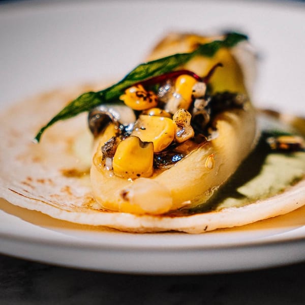 Close-up of a taco with corn, mushrooms, and a green leaf on a white plate.