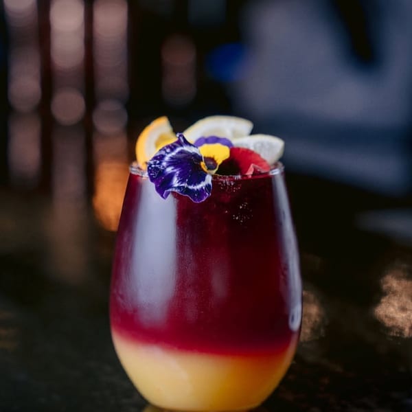 Layered red and yellow cocktail garnished with citrus slices and edible flowers on a dark surface.