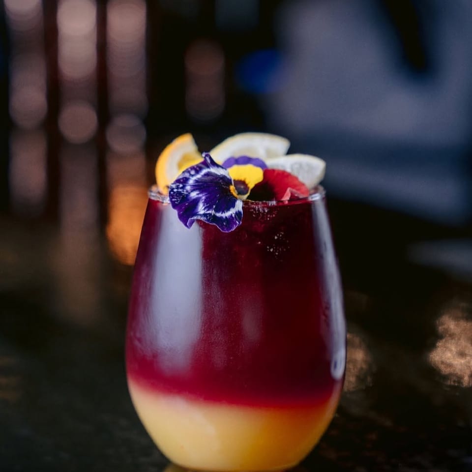 Layered red and yellow cocktail garnished with citrus slices and edible flowers on a dark surface.