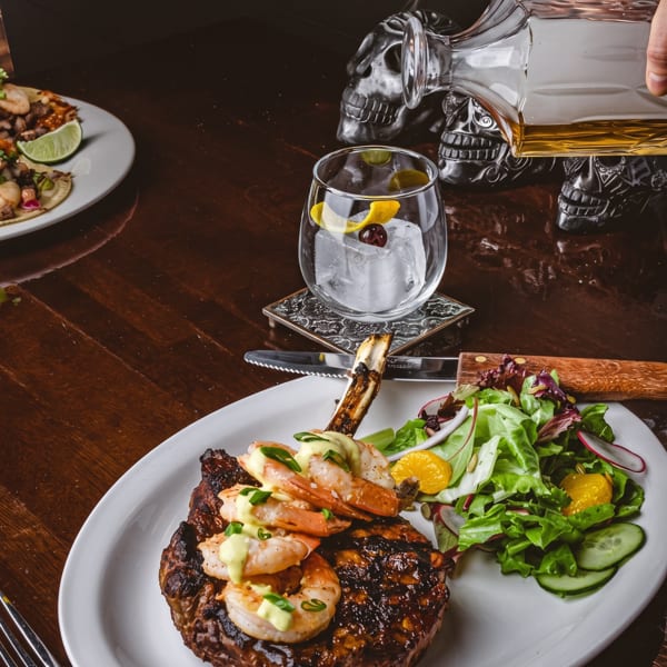 Grilled steak with shrimp and salad, a cocktail, and tacos on a dark wooden table.