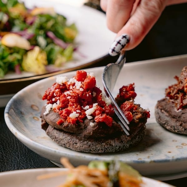 A hand with painted nails uses a fork to cut into a red and white topped appetizer.