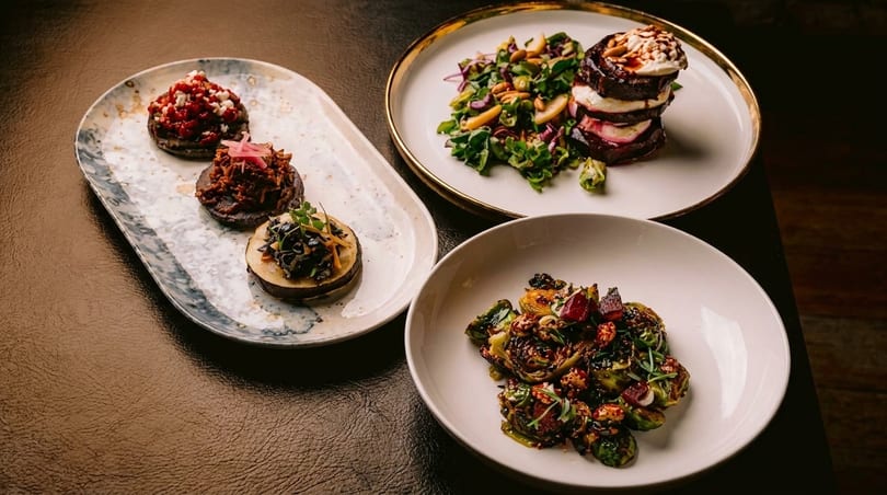 Three plates of gourmet dishes, including beet and goat cheese stacks, and roasted Brussels sprouts.